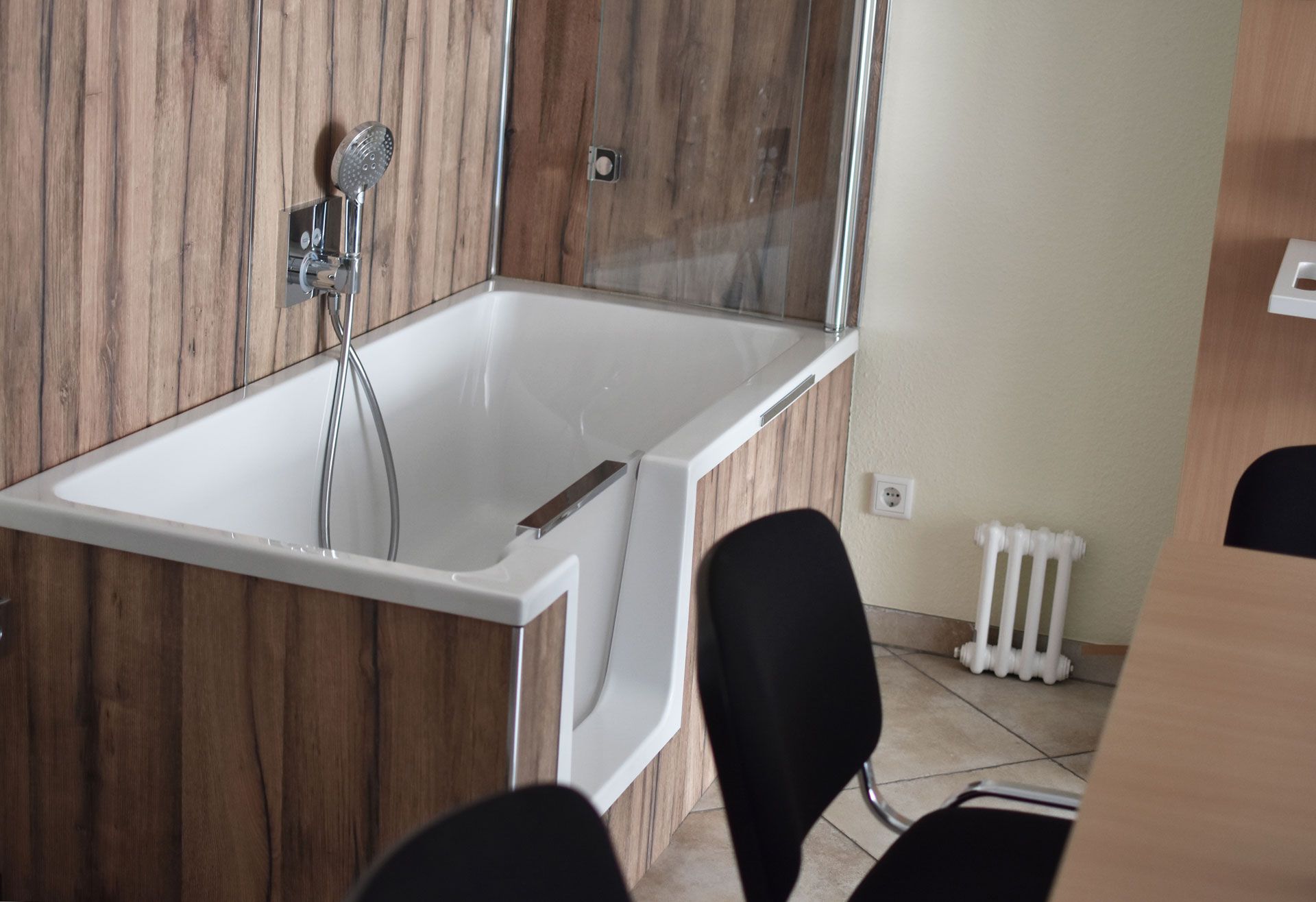Sitzbadewanne mit Duschfunktion und Holzverkleidung in einem Beratungsraum mit Stühlen und Fliesenboden.